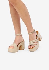 Sandalias de plataforma beige con textura tejida. Presentan correas entrecruzadas y hebilla en el tobillo. Tacón elevado con un acabado de madera natural.