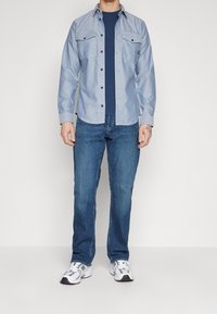 Hombre con camisa de botones azul claro sobre una camiseta azul marino, jeans azules y zapatillas blancas, de pie frente a un fondo blanco.
