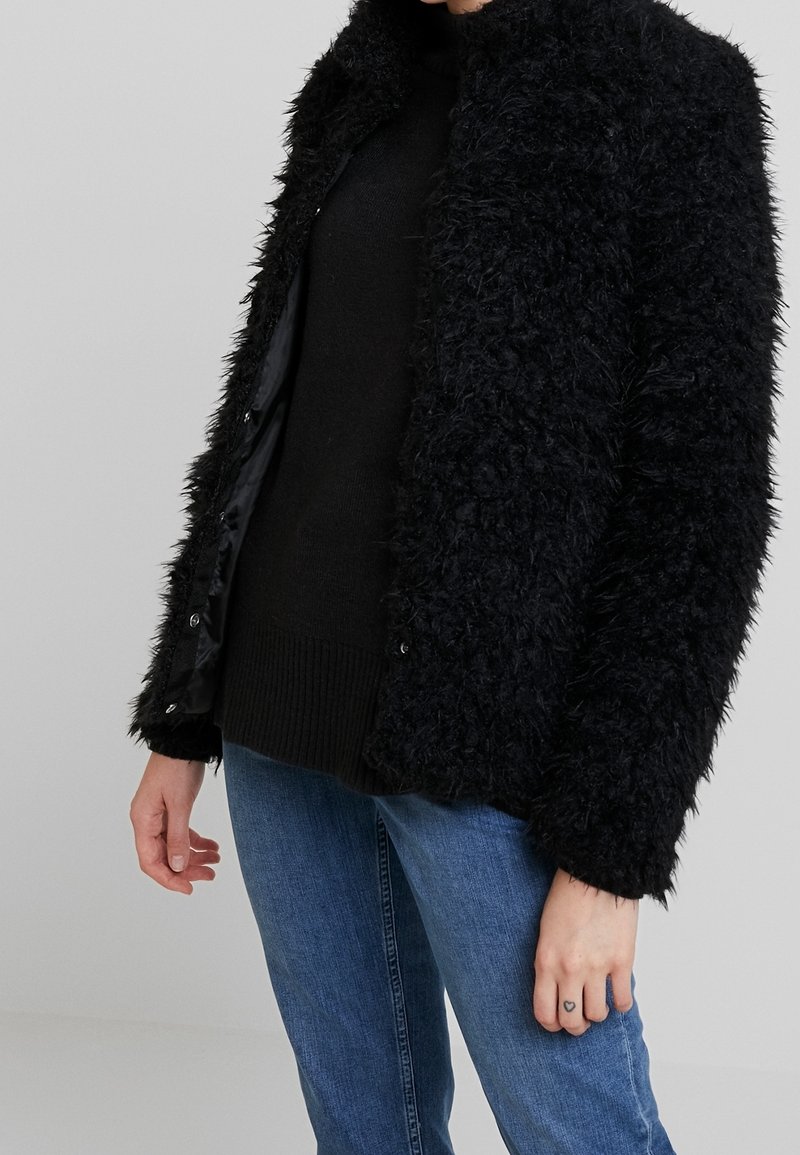 Veste en fausse fourrure noire avec un col haut, texture douce et fermeture à boutons, associée à un pull noir et un jean bleu.