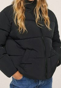 Czarna kurtka puchowa z wysokim kołnierzem, zamek błyskawiczny z przodu i kieszeniami po bokach. Posiada pikowane sekcje oraz miękką, gładką fakturę.