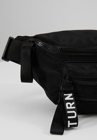 Sac banane noir avec sangle réglable, deux compartiments zippés et une languette noire étiquetée "TURN" en texte blanc.