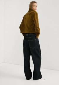 Mujer con una chaqueta corta de ante marrón y jeans negros de pierna ancha, de pie y mirando por encima del hombro contra un fondo blanco liso.