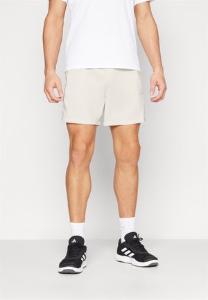 Beige Sportshorts mit elastischem Bund, versehen mit einem hellvioletten Adidas-Logo. Kombiniert mit schwarzen Turnschuhen und weißen Socken.