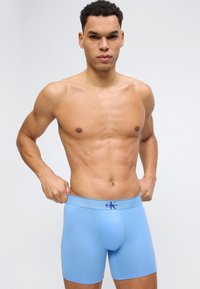 Bar overkropp, mann som justerer lyseblå Calvin Klein boxershorts, stående mot ensfarget lys grå bakgrunn.