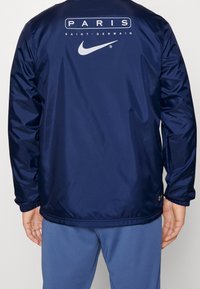 Navy nylon jacket featuring "PARIS SAINT-GERMAIN" and Nike logos in white, elastic cuffs, and a relaxed fit.