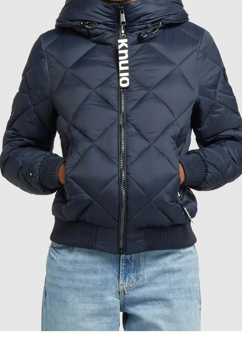 Chaqueta acolchada de estilo puffy en color marino con capucha, cierre delantero con cremallera, puños acanalados y una etiqueta de logo llamativa en la cremallera. Textura suave y lisa.