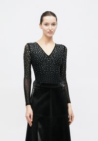 Μαύρο κορμάκι με V-λαιμόκοψη, μακριά διαφανή μανίκια και ασημένιες διακοσμητικές χάντρες. Συνδυασμένο με μαύρη φούστα από δερματίνη με υφή.