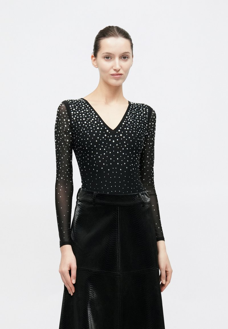 Μαύρο κορμάκι με V-λαιμόκοψη, μακριά διαφανή μανίκια και ασημένιες διακοσμητικές χάντρες. Συνδυασμένο με μαύρη φούστα από δερματίνη με υφή.