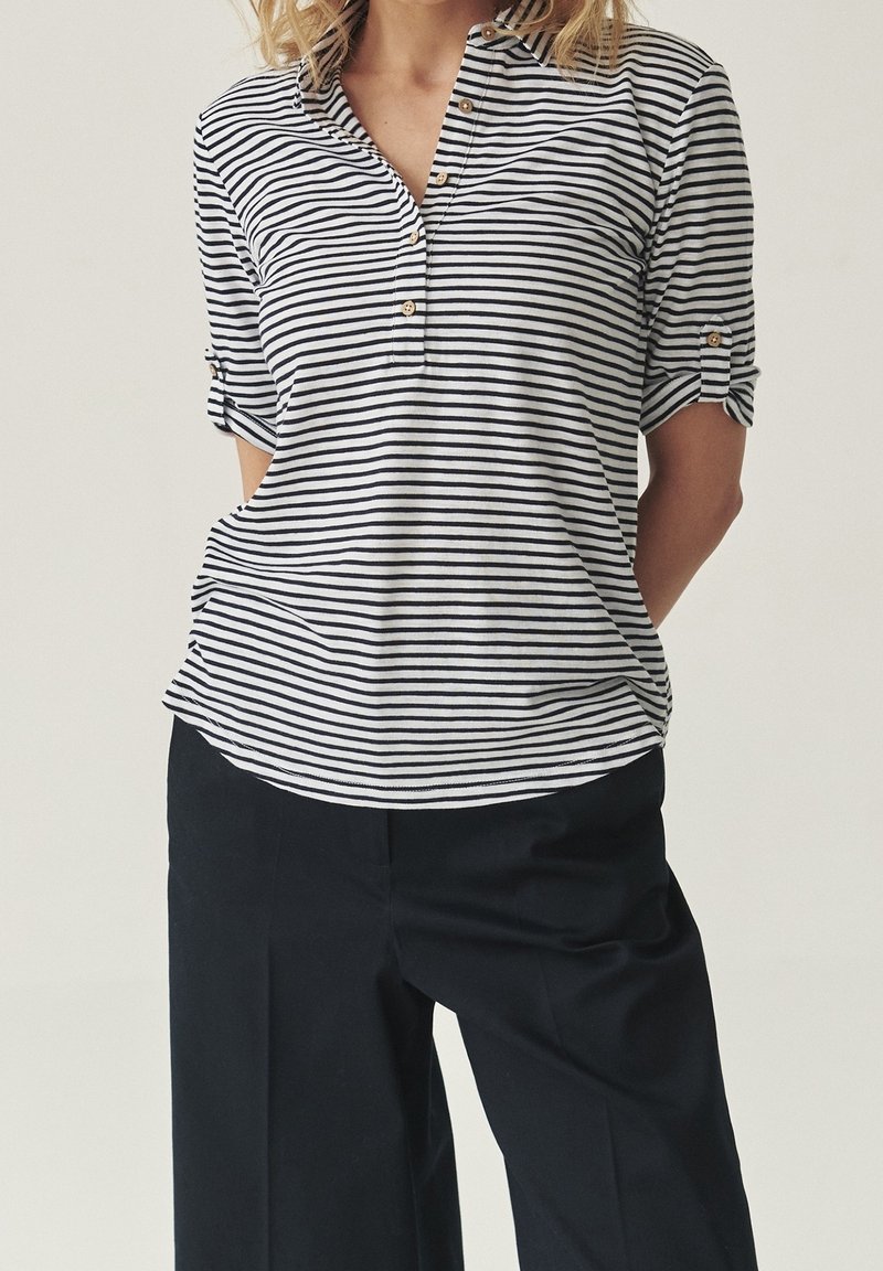 Personne portant un haut à boutons rayé noir et blanc avec manches retroussées et pantalon noir ample, debout les bras derrière le dos.