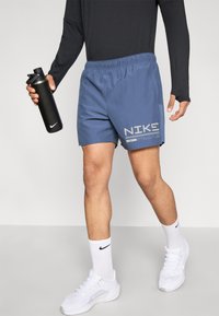 Herreblå Nike-shorts med elastisk midjebånd, med en stor hvit logo og diagonalt stripemønster. Holder en svart vannflaske i hånden.