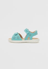 Ljusblå sandal tillverkad av syntetmaterial, med utskurna blommönster, justerbart band och vitt gummisul med texturerat grepp.