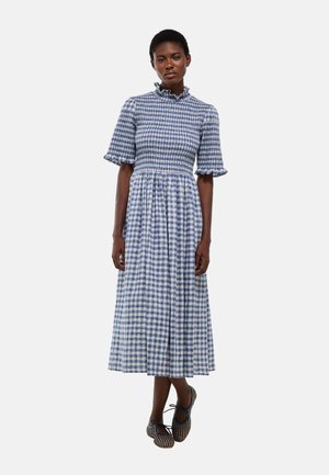 Femme portant une robe midi à carreaux bleus et blancs avec des manches courtes volantées et des ballerines noires à motifs, debout sur un fond uni.