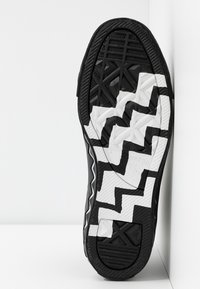 Semelle de basket noir et blanc avec motif en zigzag et prise texturée, positionnée verticalement contre un coin blanc en arrière-plan.