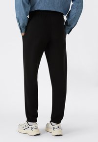 Persona vistiendo pantalones de chándal negros, camisa de mezclilla azul, calcetines blancos y zapatillas blancas, de pie con las manos en los bolsillos, de espaldas.