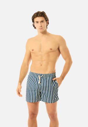 Oberkörperfreier Mann steht mit einer Hand in der Tasche, trägt blau-weiß vertikal gestreifte Badeshorts mit weißem Kordelzug.