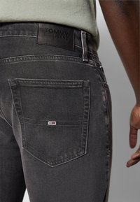 Jeans de mezclilla negra con un acabado texturizado. Presenta una etiqueta de cuero con "TOMMY JEANS" y un pequeño logo en el bolsillo trasero. Diseño clásico de cinco bolsillos.