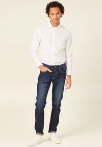 Witte lange mouwen blouse, donkerblauwe jeans en witte sneakers. Model staat tegen een neutrale achtergrond met de handen in de zakken.