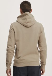 Beige hoodie med snøre i hætten, ribbede manchetter og søm, blød tekstur og et enkelt design. Bæres fra ryggen, som viser en ren finish.