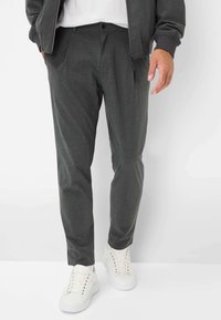 Donkergrijze op maat gemaakte broek met een gladde textuur en voorplooien, gecombineerd met witte lage sneakers. Subtiele stikdetails zijn zichtbaar.