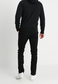 Sweatshirt à capuche noir avec une texture lisse, associé à un pantalon noir slim et des baskets blanches ; design minimaliste, sans motifs visibles.