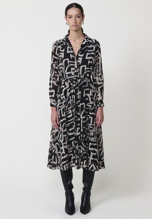 Mujer vestida con un vestido midi de patrones abstractos en negro y beige, con mangas largas y botas altas negras, de pie contra un fondo blanco.