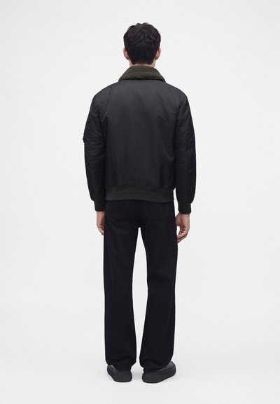 Homme portant une veste bomber noire avec un col en shearling vert foncé, un pantalon noir et des chaussures noires, debout de dos sur un fond blanc.