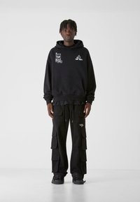 Fekete kapucnis pulóver grafikus díszítéssel és elülső zsebbel, fekete cargo nadrággal párosítva, amely több zsebbel rendelkezik, mindkettő laza szabású.