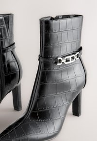 Black faux crocodile leather ankle boots with pointed toes, block heels, side zippers, and silver chain detail around the ankles.