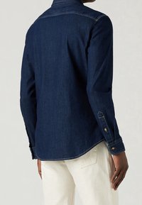 Camicia in denim blu scuro con maniche lunghe, colletto e bottoni decorativi sui polsini, con orlo cucito e vestibilità rilassata.