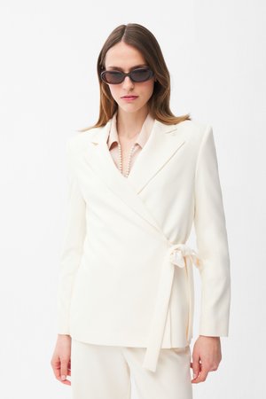 Mujer con blazer blanco cruzado atado al lateral, pantalones blancos, collar de perlas y gafas de sol oscuras, sobre fondo blanco liso.