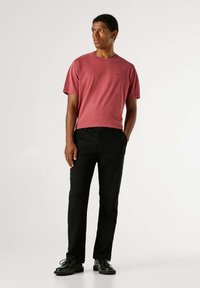 Rode katoenen T-shirt met korte mouwen en een ronde hals, gecombineerd met een zwarte broek en zwarte schoenen, weergegeven tegen een effen achtergrond.