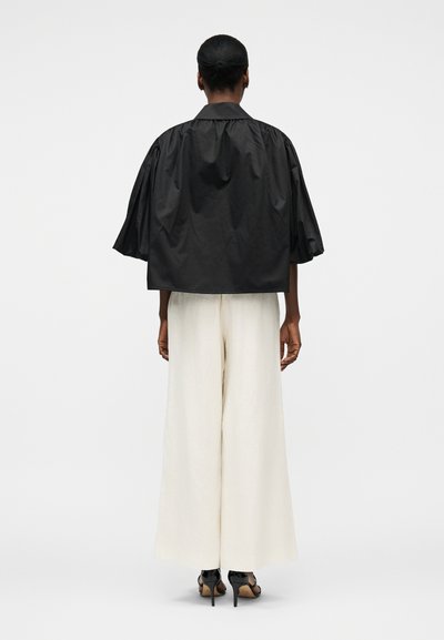 Femme aux cheveux courts portant une veste noire ample à manches courtes, un pantalon blanc à jambes larges et des chaussures à talons noirs, debout de dos.