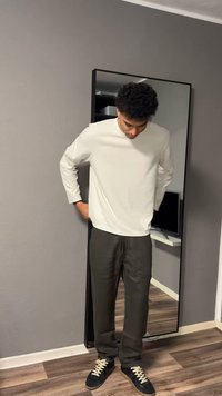 Άνδρας με λευκό πουκάμισο μακρυμάνικο και σκουρόχρωμο παντελόνι που ρυθμίζει τα ρούχα του μπροστά σε καθρέφτη ολόσωμο σε δωμάτιο με γκρι τοίχους.