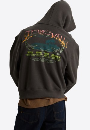Person, die einen dunklen Kapuzenpullover mit buntem „Off The Wall“-Text und Grafik von Wellen und Surfspots auf dem Rücken trägt.