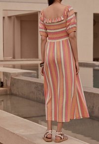 Femme se tenant près d'une mare peu profonde portant une robe midi ray ée pastel avec des manches bouffantes et des sandales gladiateur blanches.