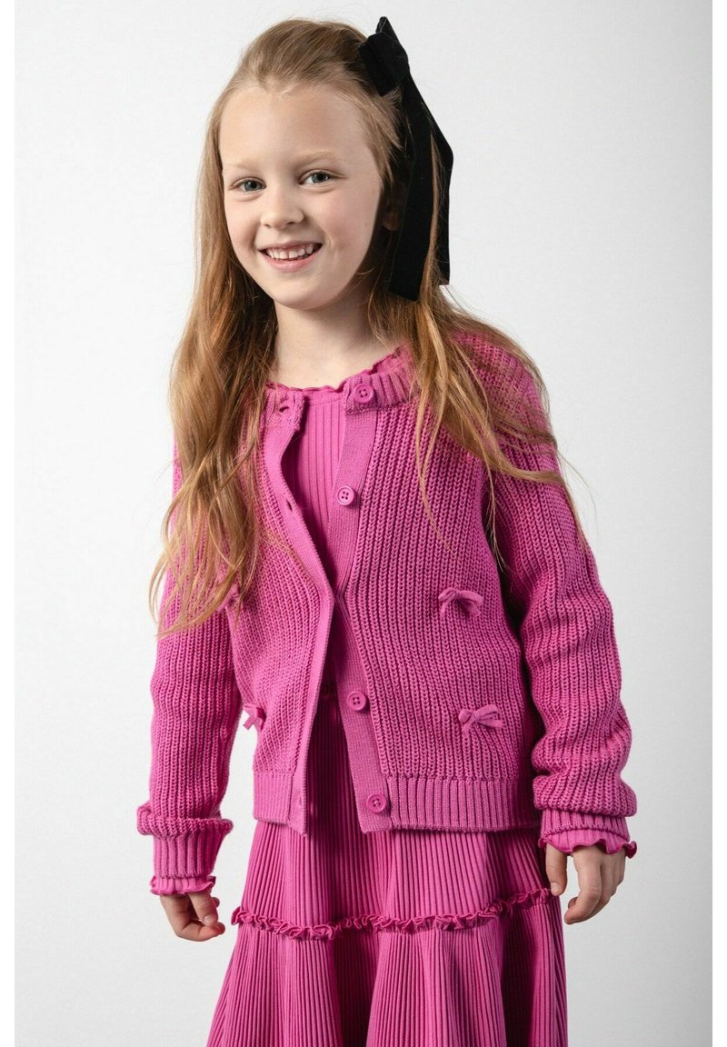 Jasnoróżowy sweter z dzianiny z zapięciem na guziki i akcentami w postaci kokardek, noszony na pasującej plisowanej sukience, prezentującej teksturowane wykończenie.