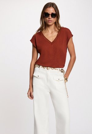 Femme portant un haut manches courtes rouge foncé à col en V, un pantalon blanc taille haute avec une ceinture chaîne dorée, et des lunettes de soleil sombres, debout avec une main dans la poche.