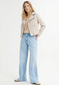 Giacca da moto in pelle beige chiaro con chiusura a zip, abbinata a jeans a gamba larga blu chiaro. Le texture contrastano delicatamente, con una finitura liscia.