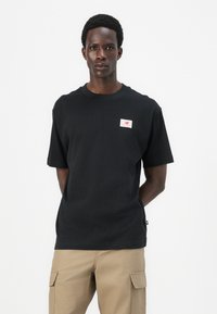 T-shirt en coton noir à manches courtes et col rond, avec un petit patch logo sur la poitrine gauche. Texture lisse et coupe décontractée.
