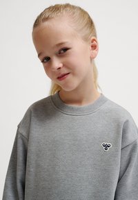 Grauer Pullover mit Rundhalsausschnitt und überschnittenen Schultern. Besitzt ein kleines gesticktes Bienenmotiv auf der Brust. Weicher, strukturierter Stoff.