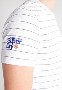 Bílá košile s krátkým rukávem a tenkými námořnickými horizontálními pruhy, se modrou vyšitou značkou "Super Dry" na levém rukávu.