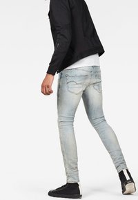 Jeans skinny de denim azul claro con desgastes, diseño de cinco bolsillos, combinados con una chaqueta negra con cremallera y zapatillas negras.