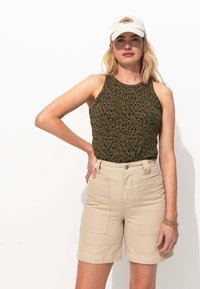 Femme portant une casquette blanche, un haut sans manches à imprimé léopard, un short beige taille haute et des bracelets en or au poignet droit.