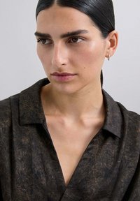 Camisa con un tejido marrón oscuro texturizado que presenta un patrón sutil. Cuello con muesca y un corte relajado. Acentos de hardware suaves y mínimos.