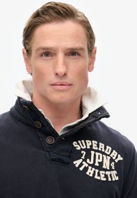 Marineblauer Pullover mit geknöpftem Kragen, ausgestattet mit weißem Fleece-Futter und gesticktem "Superdry JPN Athletic"-Design auf der Vorderseite.