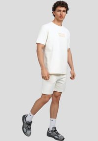 Camiseta de algodón blanca con la impresión "WRONG FRIENDS", combinada con pantalones cortos blancos y zapatillas deportivas grises y negras, contra un fondo neutro.