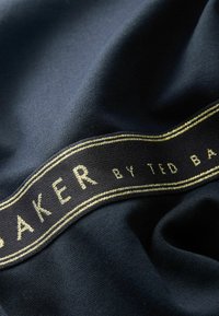 Un tissu bleu marine au toucher lisse présente une étiquette noire avec des lettres dorées indiquant "BAKER by TED BAKER", mettant en valeur des détails de design élégants.