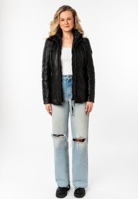 Vrouw met blond haar, gekleed in een zwart leren jack, witte top, gescheurde lichtblauwe jeans en zwarte stevige schoenen, staand tegen een witte achtergrond.