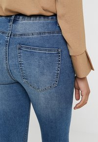 Vaqueros ajustados de mezclilla azul con cintura de media altura, que cuentan con un único bolsillo trasero y costuras en contraste. La tela tiene una textura suave y desgastada.