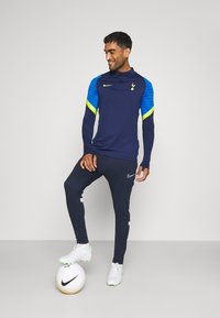 Marine atletische set met blauwe en neongele accenten; lange mouwen top met een halve ritssluiting en logo, slim-fit broek gecombineerd met witte voetbalschoenen en een bal.